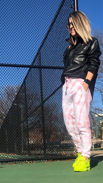 Woman wearing sunglasses, leather jacket, yellow sneakers, and pink tie dye joggers standing on tennis court with hands in pockets