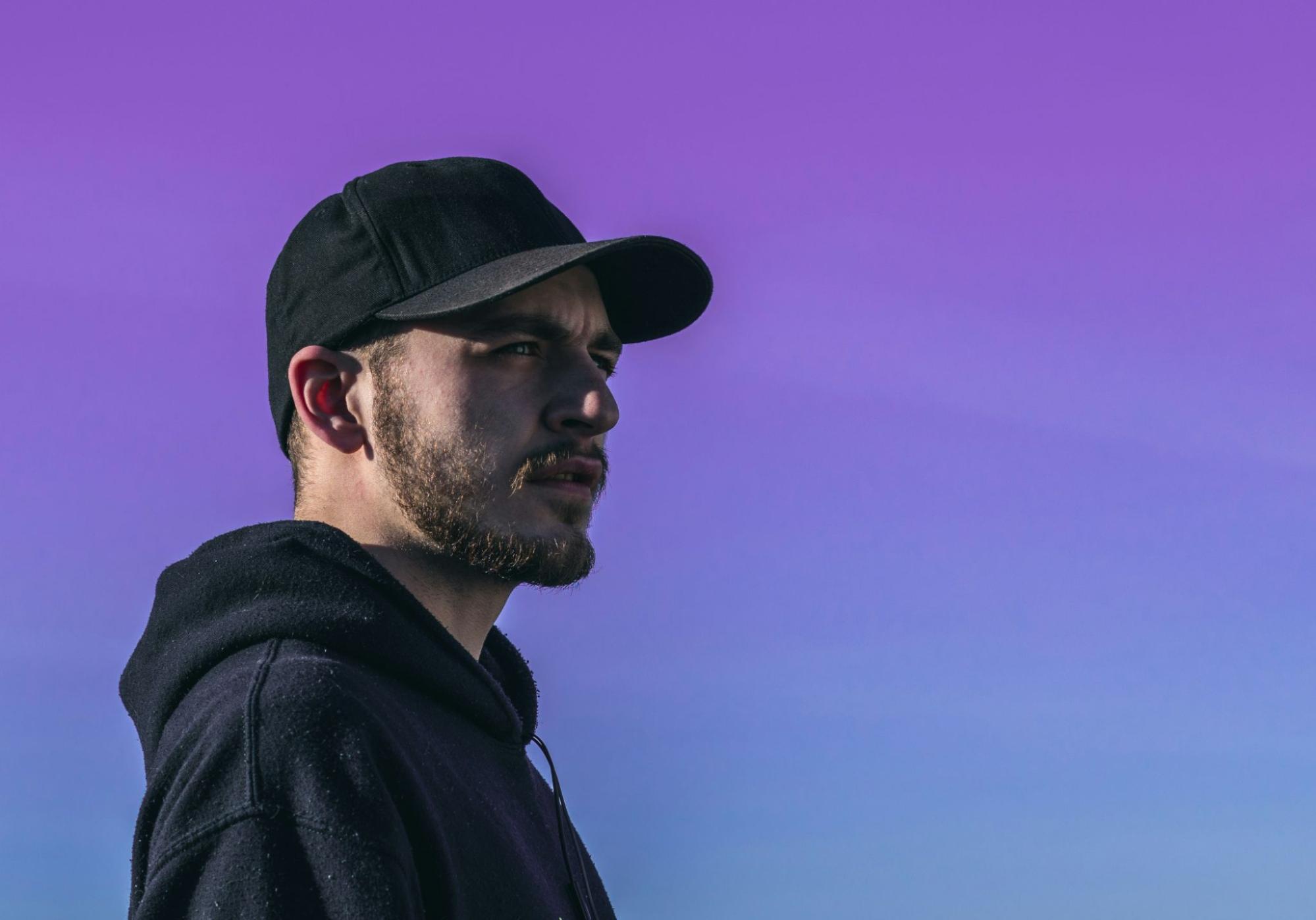 A man wearing a black fitted hat and a hoodie against a purple sky.
